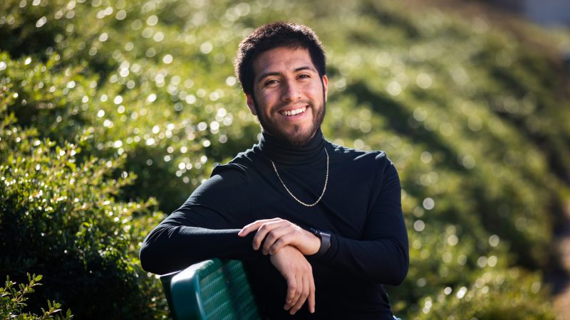 A man in a black turtleneck smiles while sitting on a bench in front of greenery