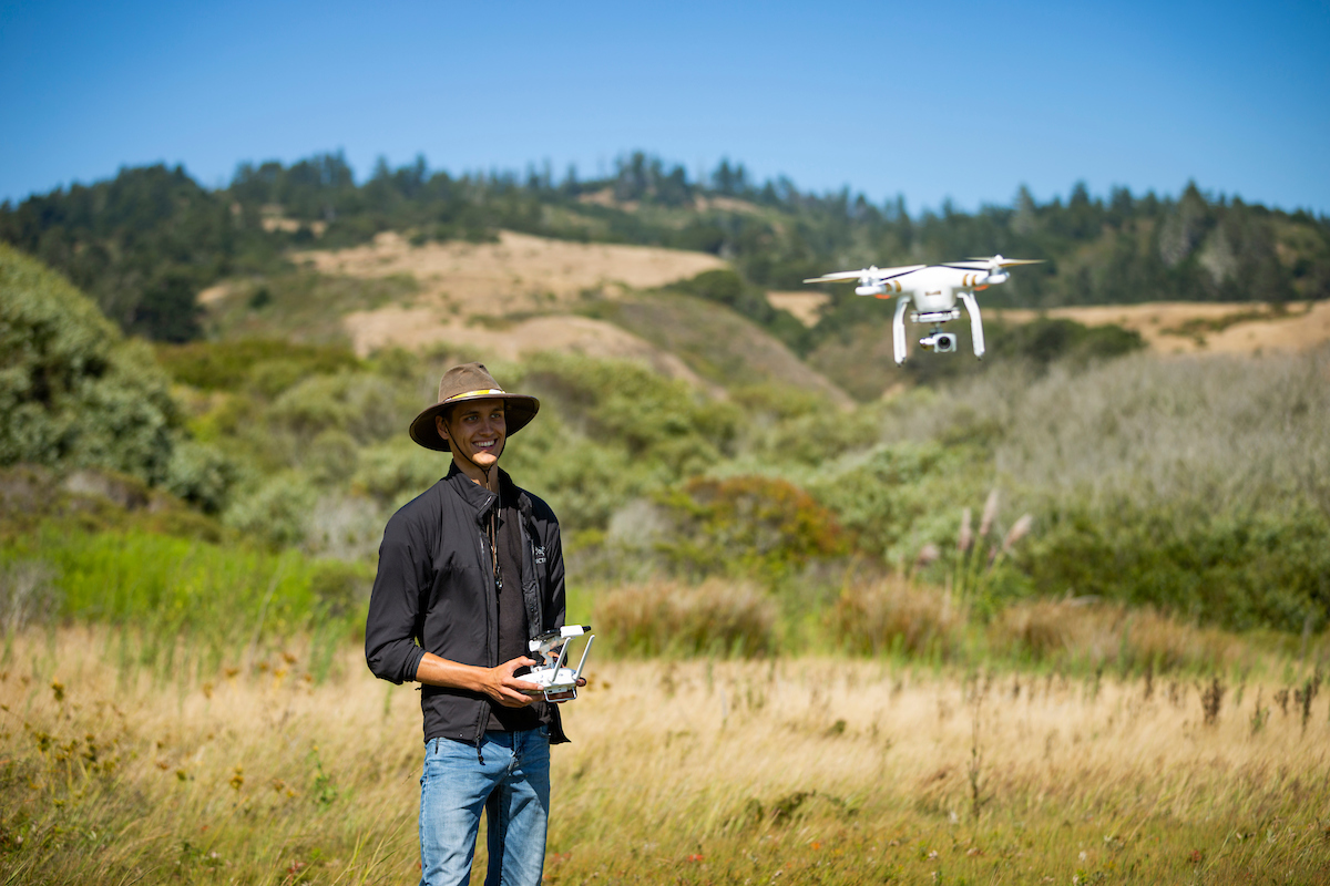 A young man in a wide brimmed hat pilots a camera drone over a grassy field on a hillside with forested mountains in the background