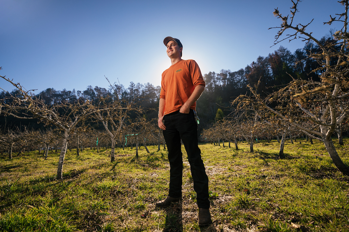 A young man in an orange shirt and baseball cap stands in an orchard with the sun behind him.