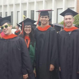 A group of 8 graduates in black robes and orange stolls smile
