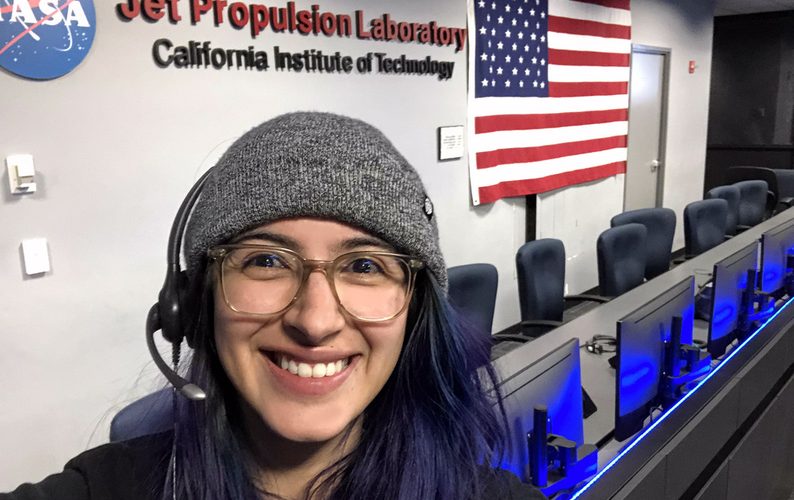 A person wearing glasses, a hat and a head set stands in the NASA Jet Propulsion Laboratory's Mission Control Room