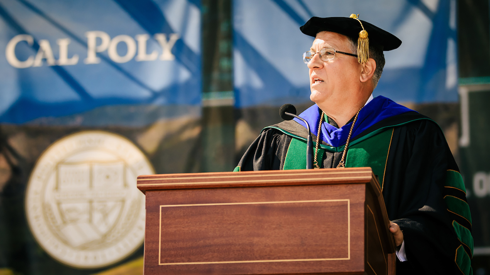 President Armstrong, in graduation regalia, stands at a podium with the sun shining on a Cal Poly banner behind him.