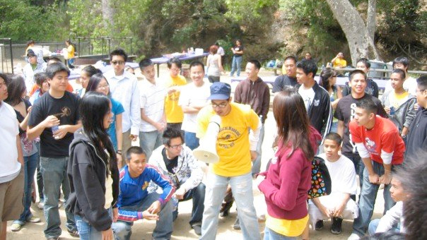 A man wearing a blue hat and yellow shirt holds a megaphone as two people play rock-paper-scissors with a crowd watching them compete