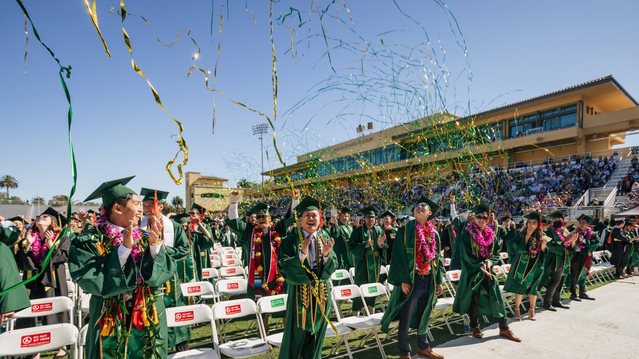 Graduates in green robes, spaced two seats apart, stand and cheer in Spanos Stadium as green and gold streamers fly through the air.