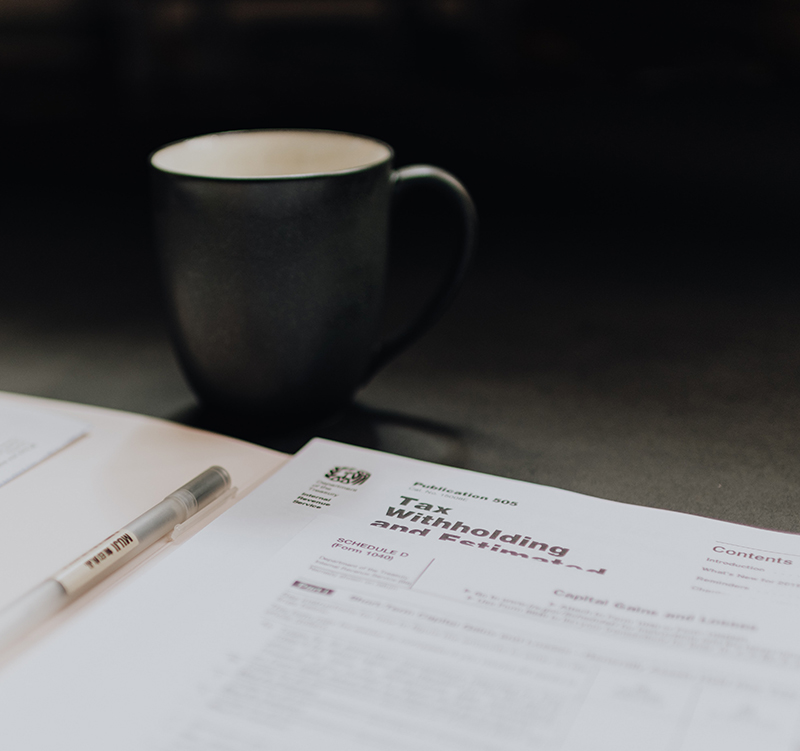 A stack of tax forms lies on a table top next to a black coffee cup and a pen