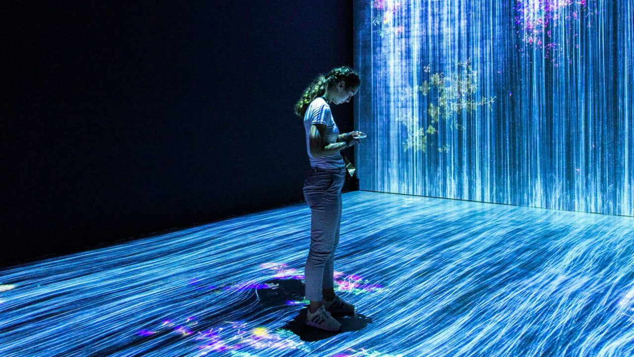 A person looking at a phone stands within an art installation with lights and blue graphics