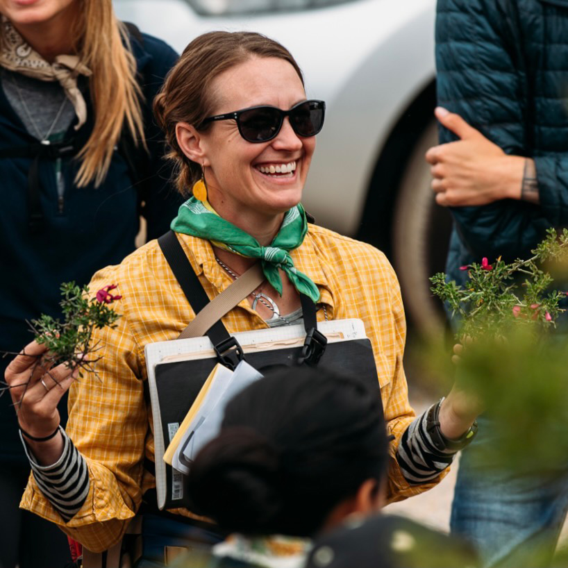 A woman wearing a yellow shirt, sunglasses and green handkerchief smiles while holding samples of plants
