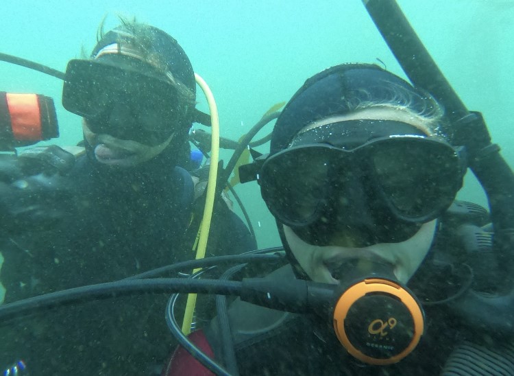 A man and woman underwater in wetsuits and SCUBA gear look into the camera