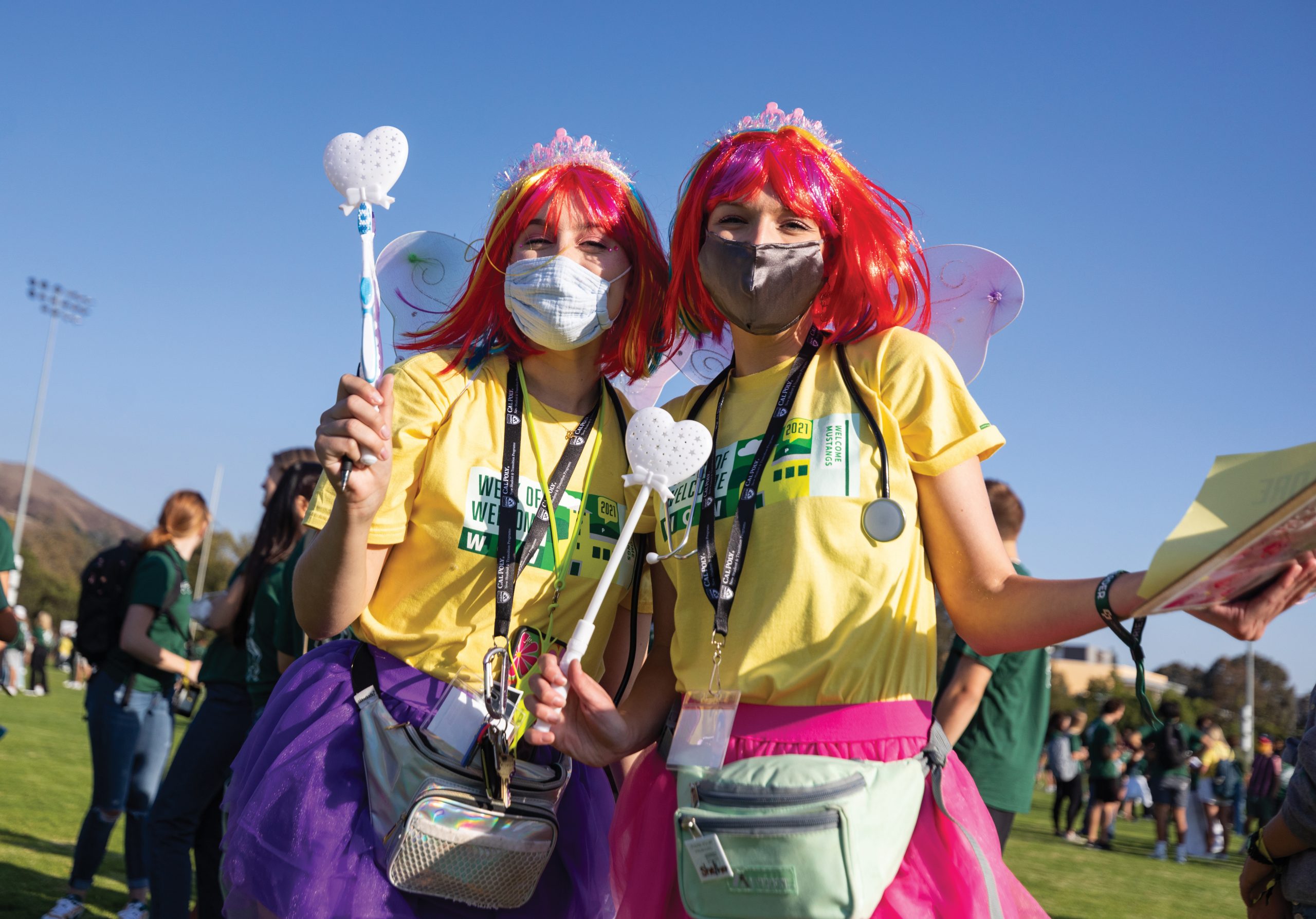 On a green field, two young women pose for a photo wearing yellow WOW leader t-shirts, bright tutus, red wigs, fairy wings and face masks.