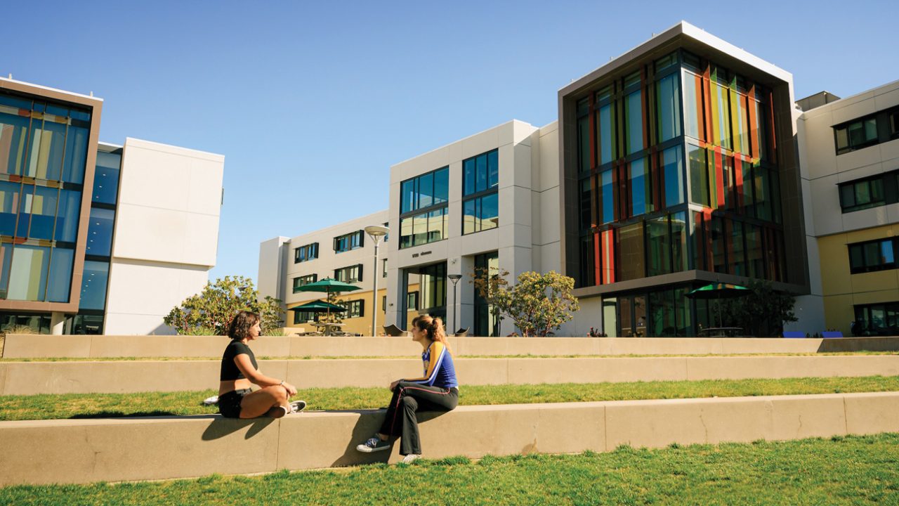 Two people sit talking in front of Cal Poly residence halls