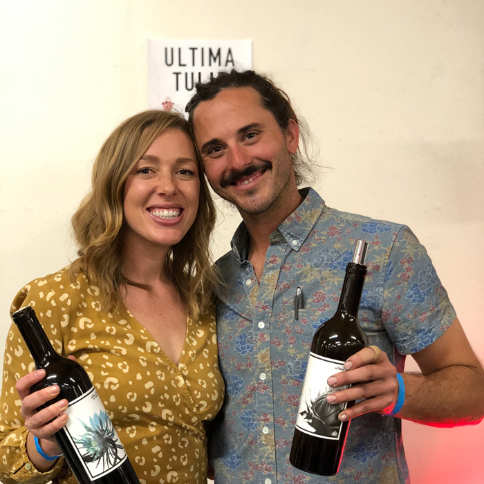 Mallory and Cameron Stoffel hold bottles of wine and smile near a sign saying 'Ultima Tulie.'