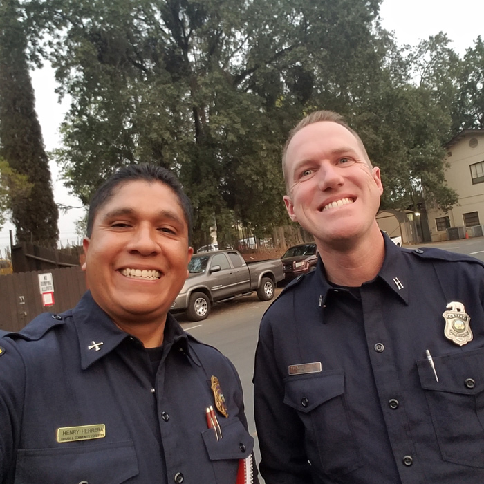 Henry Herrera and Thomas Shoots smile wearing their CAL FIRE uniforms in front of trees and cars