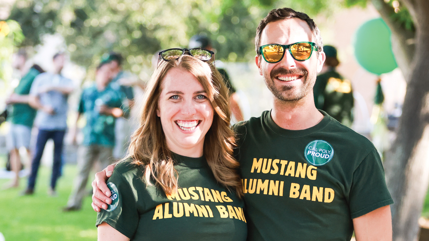 Two people smile at a tailgate party wearing shirts saying 'Mustang Alumni Band.'