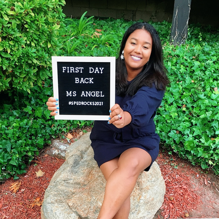 Angel Key sits on a rock and holds a sign saying 'First Day Back Ms. Angel #spedrocks2021".
