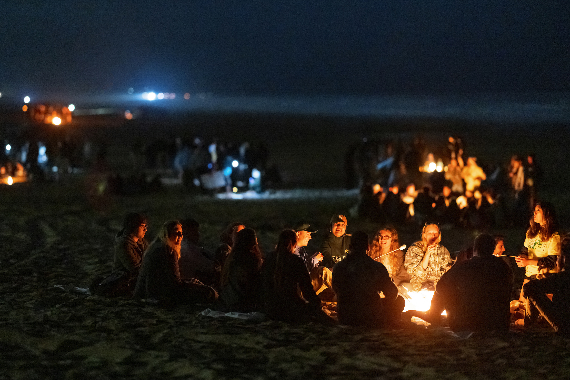 On a dark beach, clusters of students sit in circles, lit by the glow of bonfires