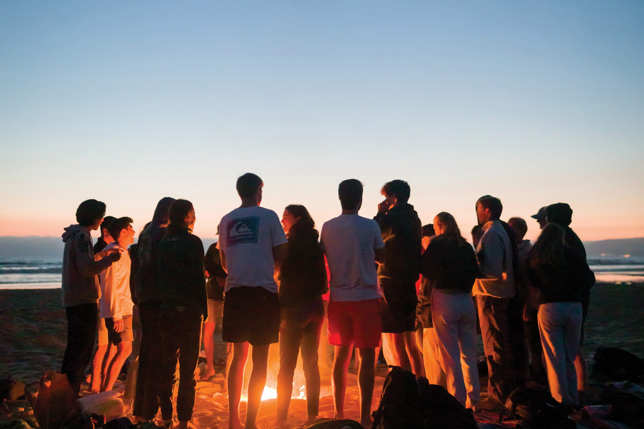 A group of students stand in a circle at sunset on the beach, lit by the glow of the bonfire at the center of the circle