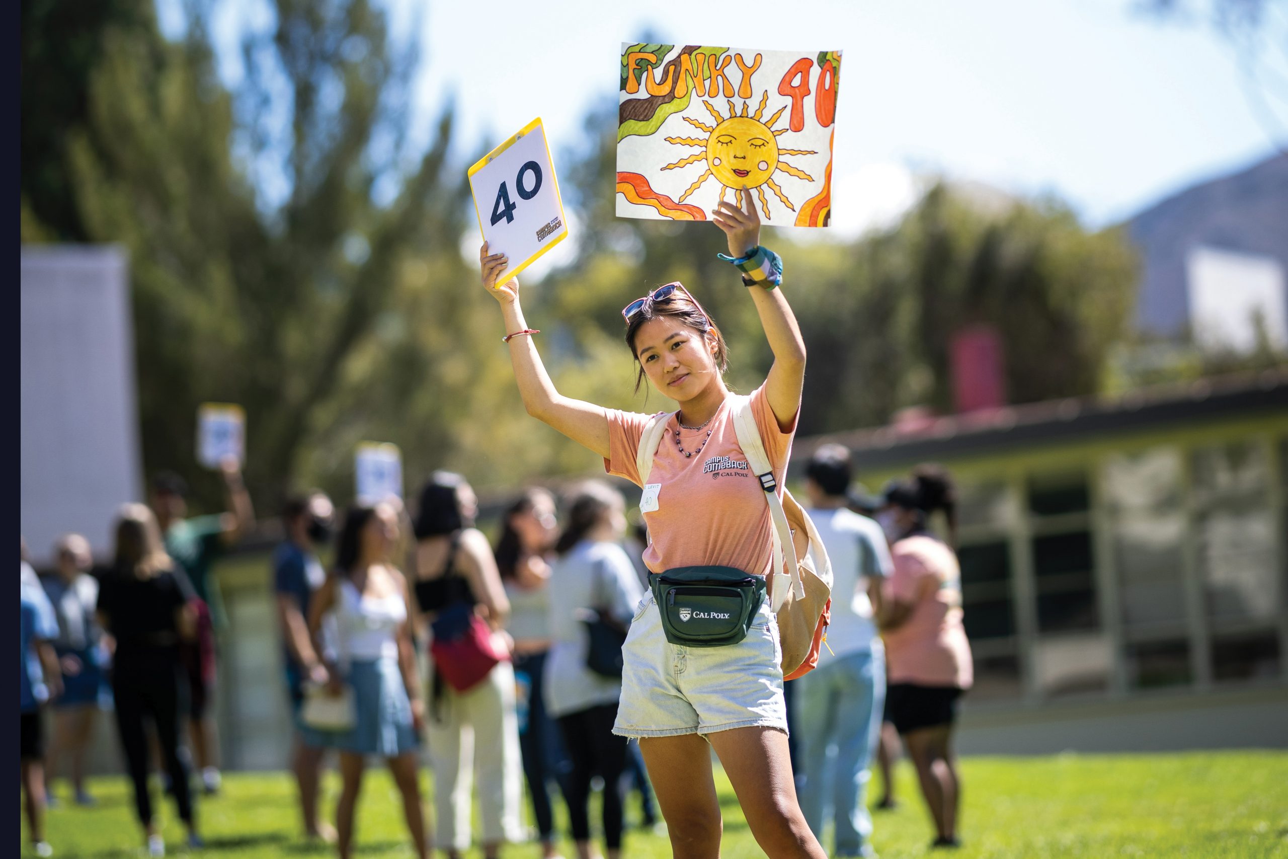 A young woman in a pink shirt holds up signs that say "40" and "funky 40" on a green campus lawn