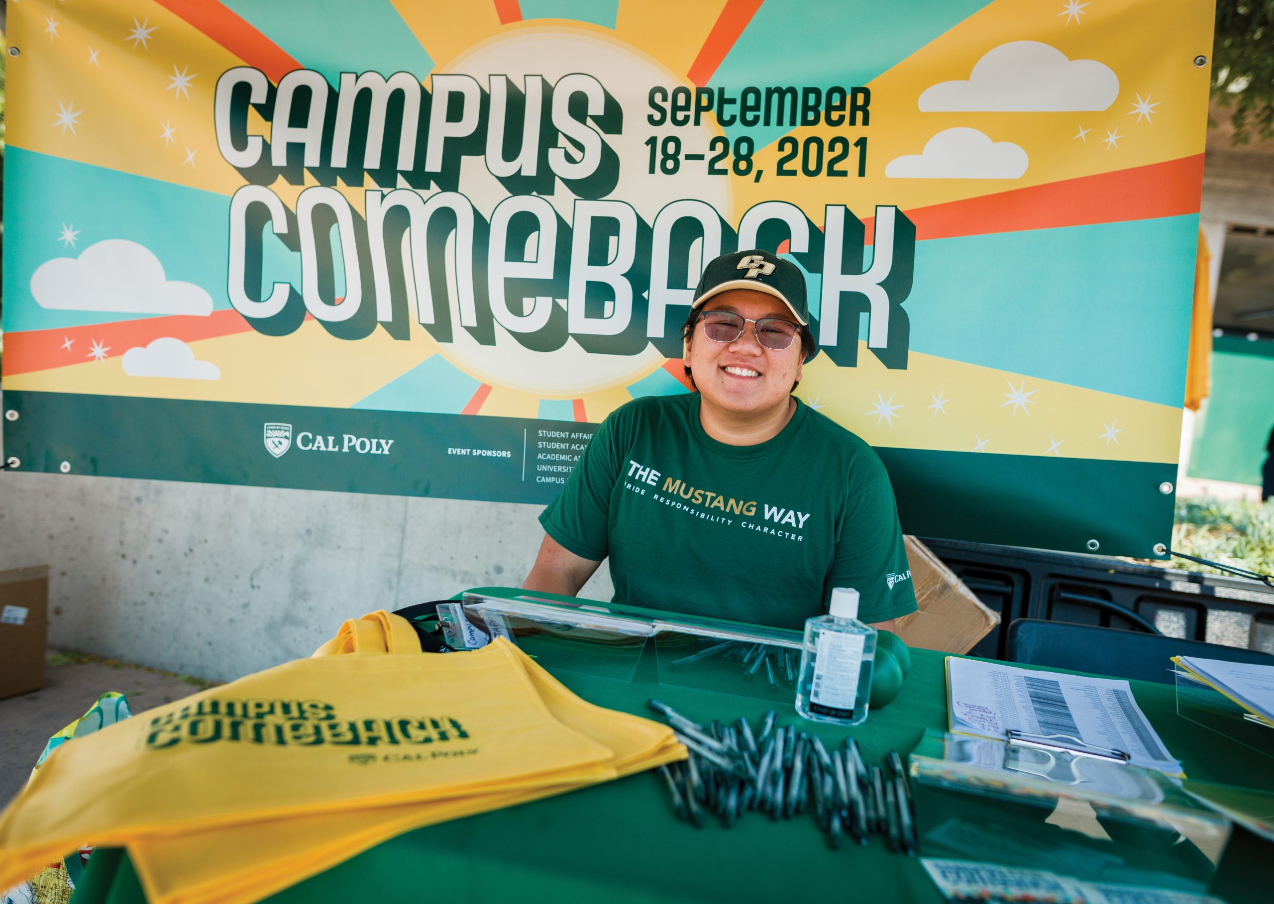 A young man in a Cal Poly t-shirt and cap sits at a table full of Cal Poly bags and pens, in front of a sign that reads "Campus Comeback"