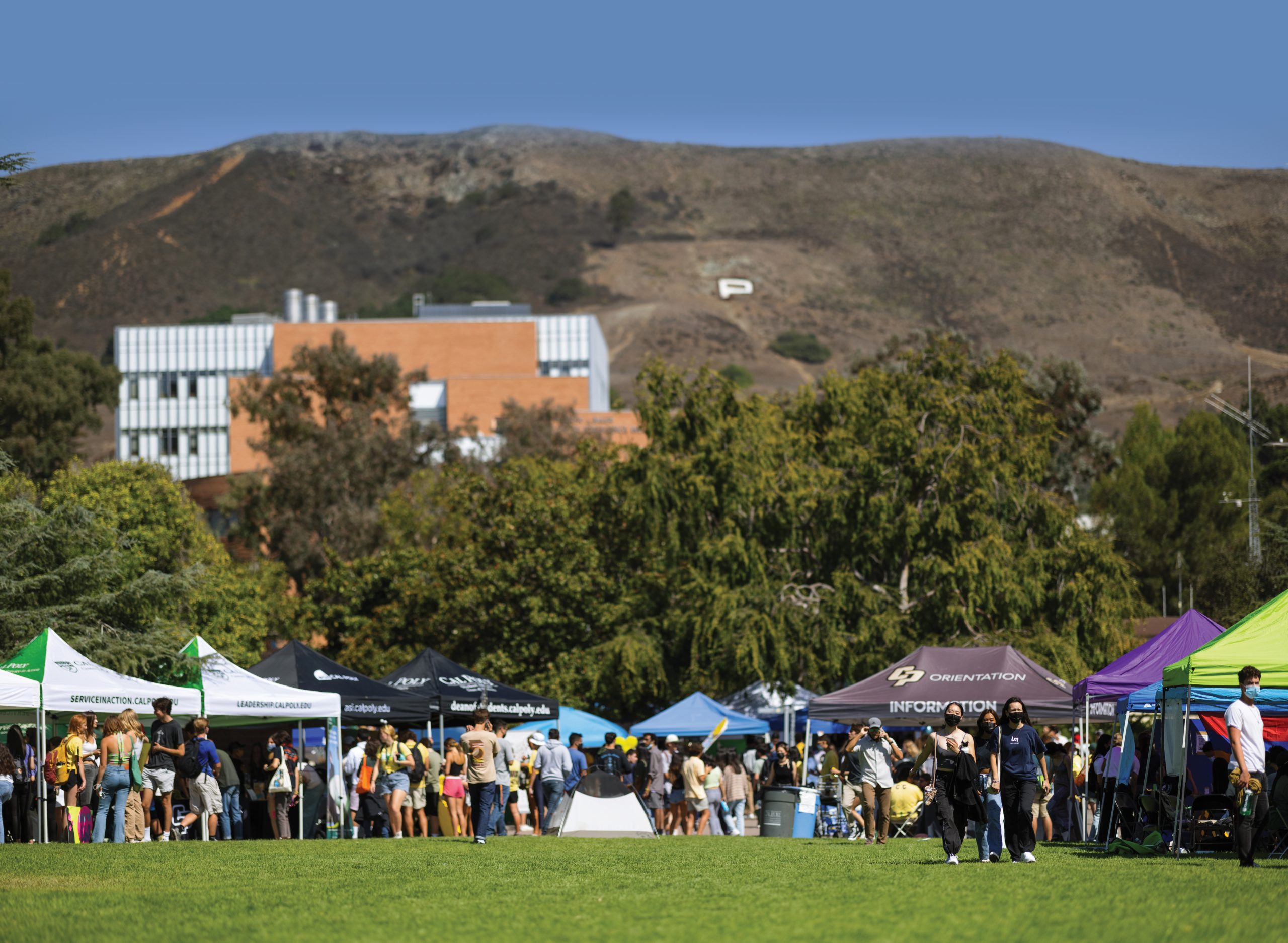 Crowds of students browse tents and booths around a green lawn, with a large metal and brick building and Cal Poly's hillside P visible in the background