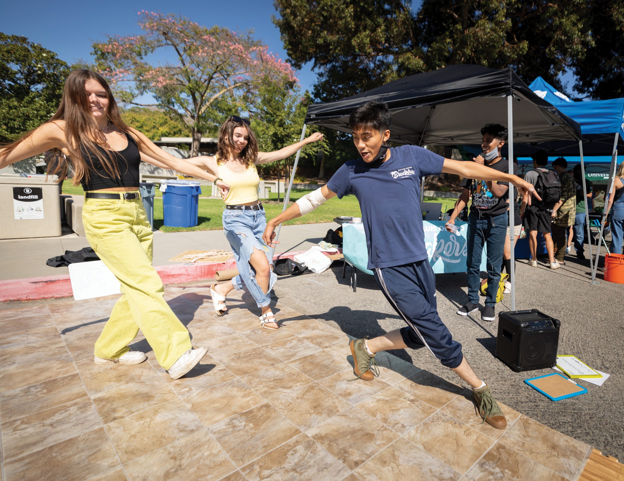In a parking lot surrounded by pop tent booths, a young man leads two young women in a synchronized breakdance move on a large sheet of linoleum