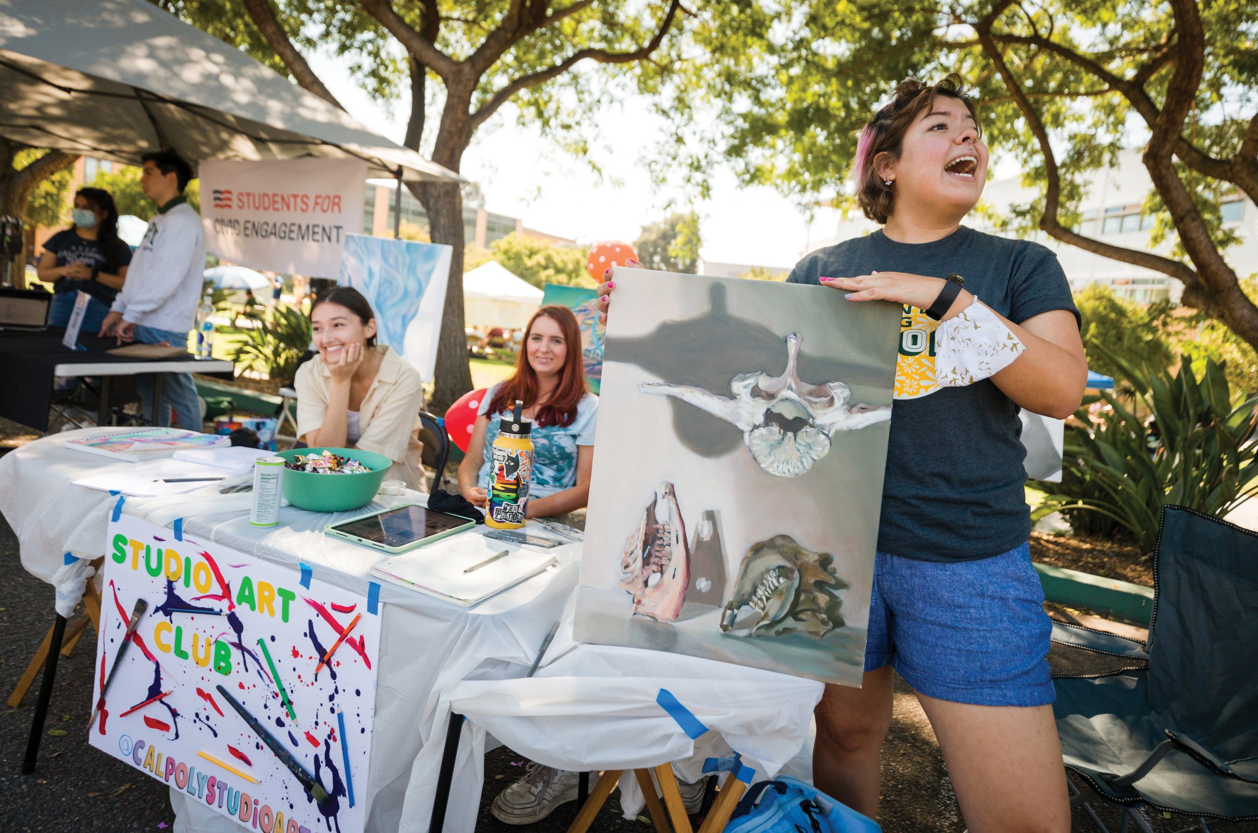 In front of a table draped with a sign reading "Studio Art Club" and splattered with paint, a young woman with pink hair highlightsholds up an abstract painting and calls to other students nearby