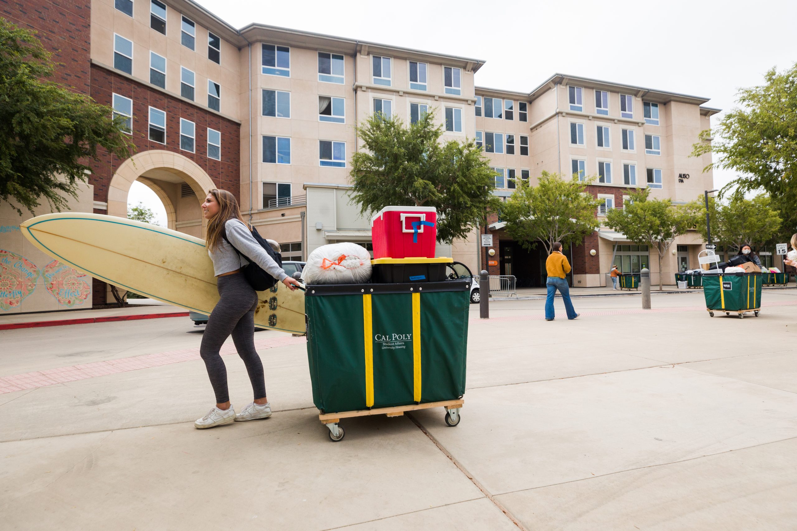 A young woman pulls a loaded moving cart with one arm and holds a surfboard tucked under the other as she walks across the courtyard at Poly Canyon Village.