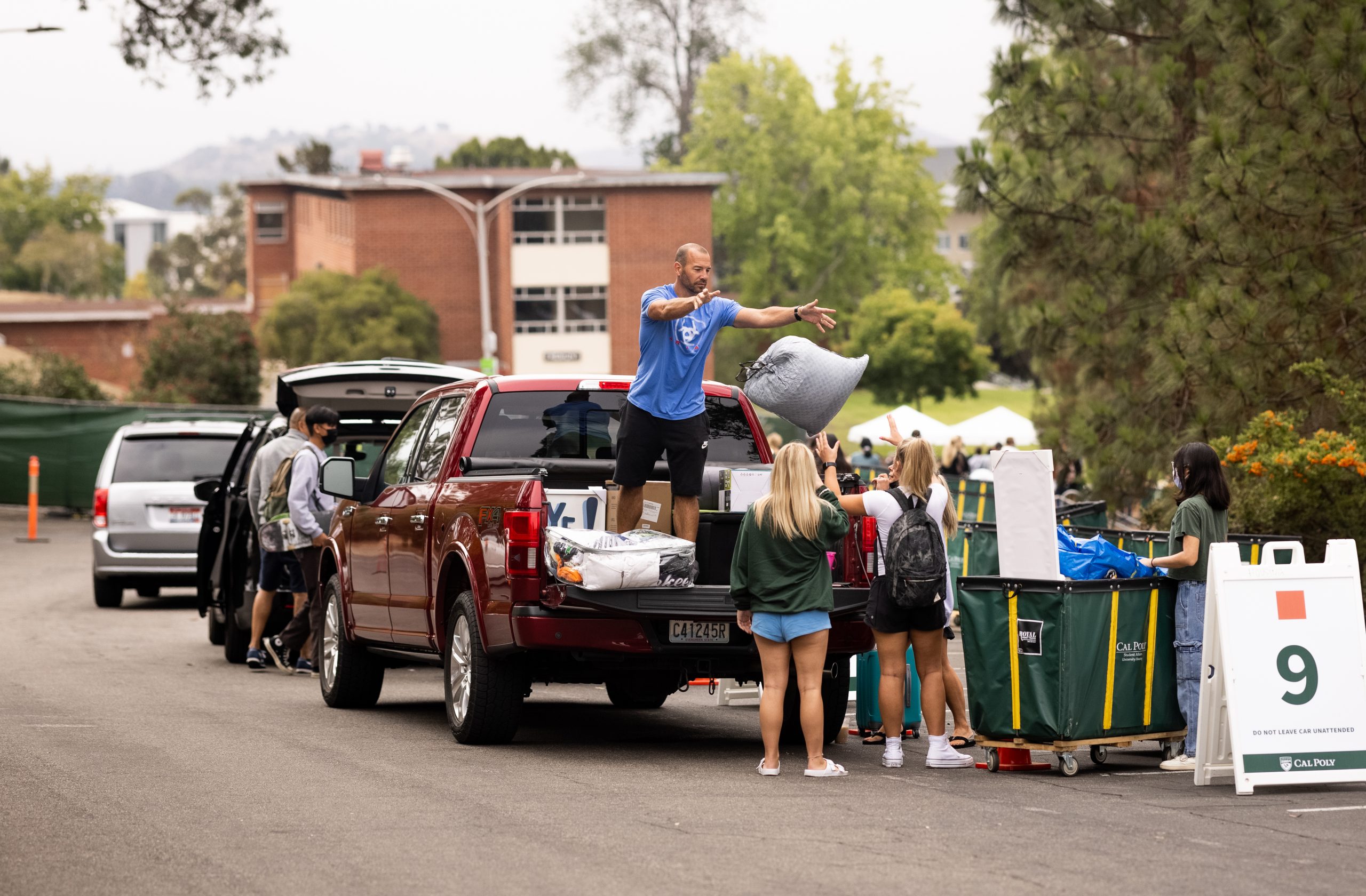 From the bed of a red truck, a man in a blue T shirt tosses a bag to a young woman wearing a backpack in front of the red brick residence halls, in a line of vehicles as families unload student belongings.