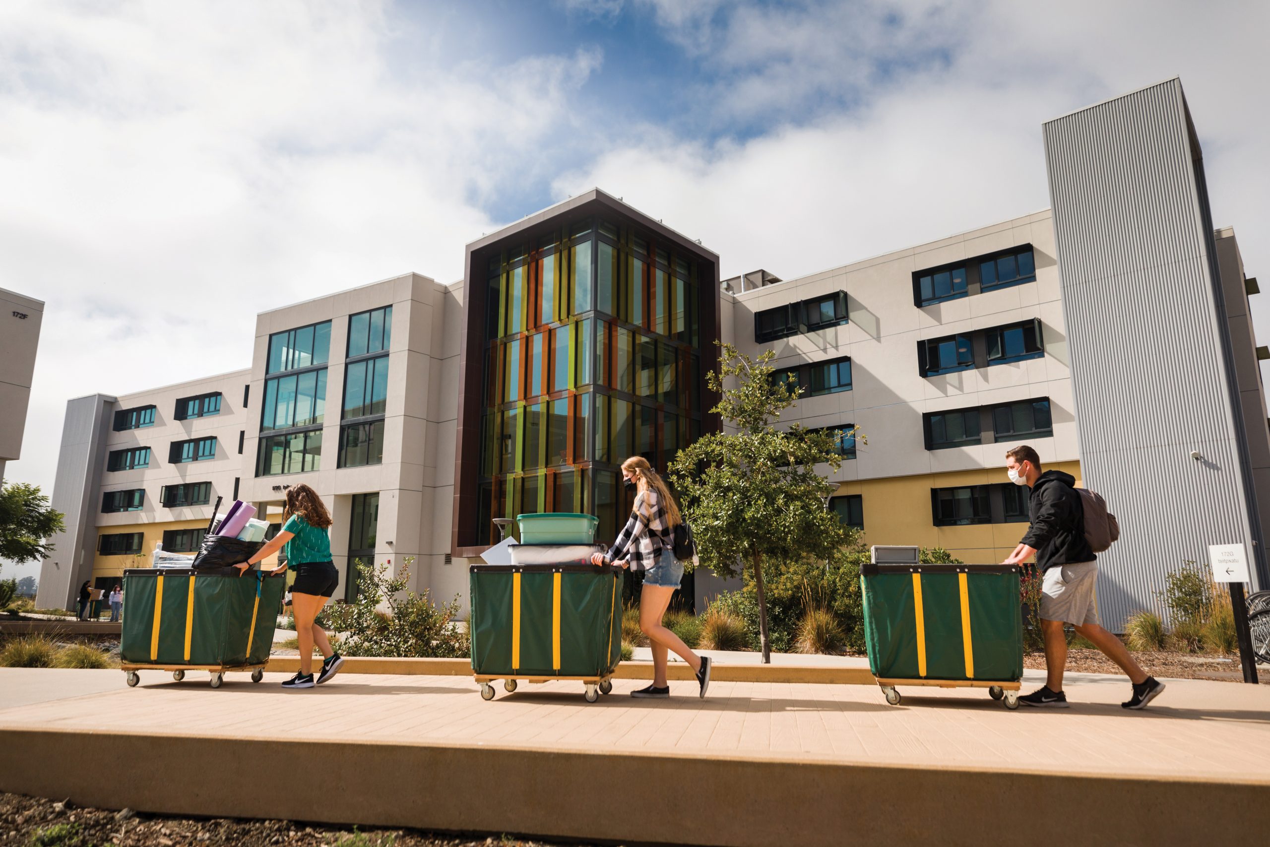 Three students push green and gold carts across a courtyard in front of a large residence hall with a multi-colored tower window