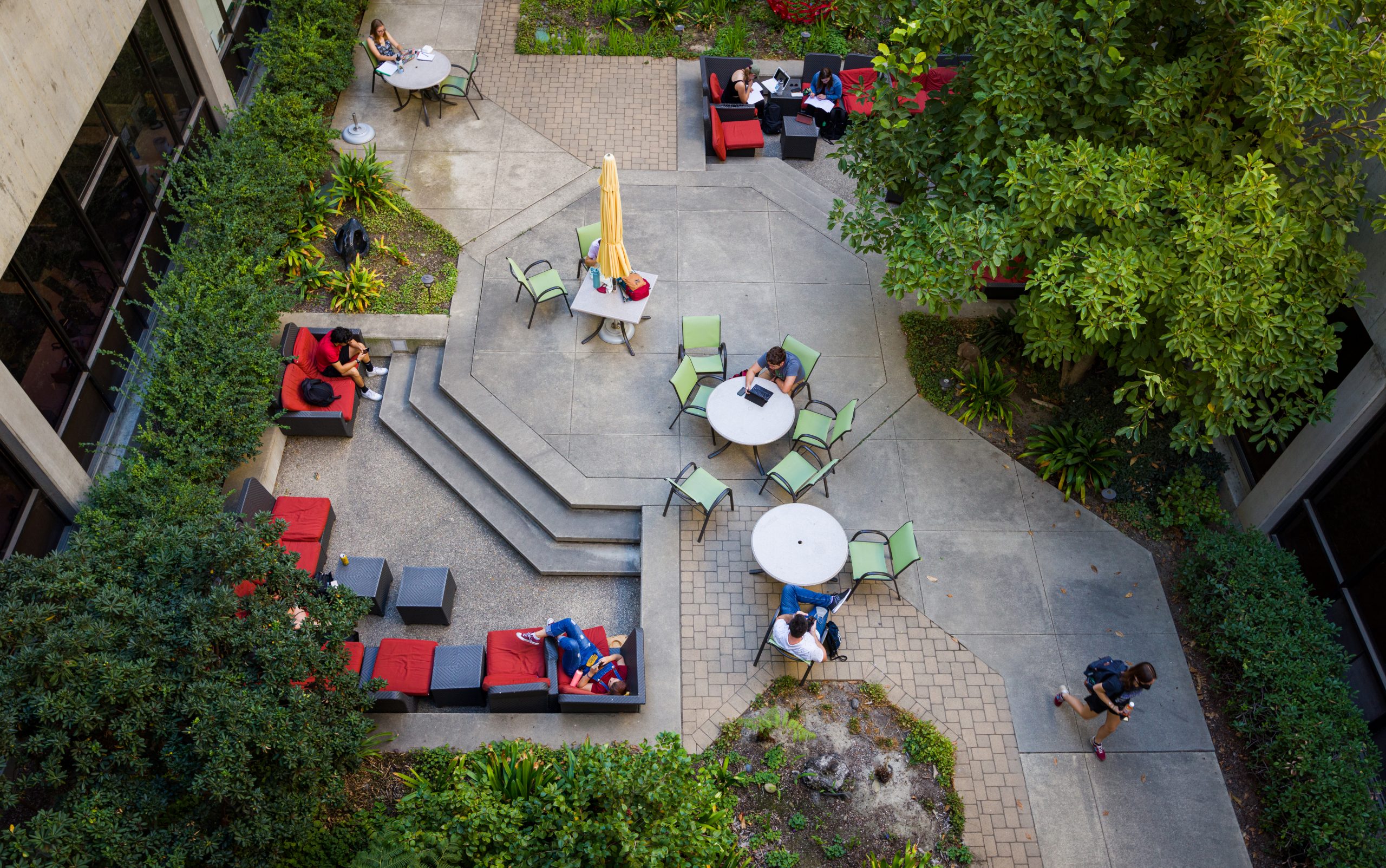Seen from above, students sit at tables and brightly-colored couches in a courtyard surrounded by trees and shrubs