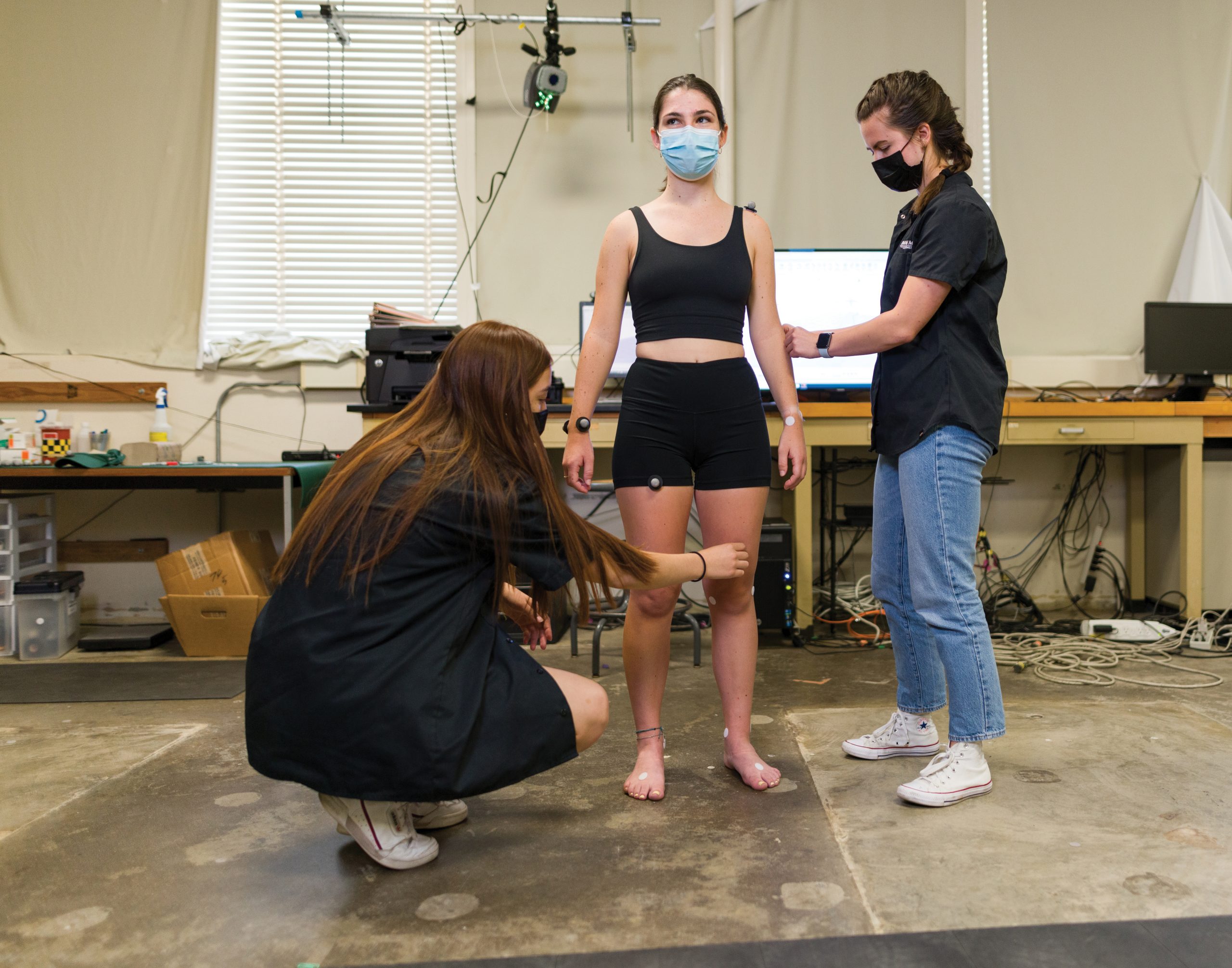 In a lab, two young women adjust sensors stuck to the arms and legs of a third, wearing close-fitting athletic clothes. All wear face coverings.