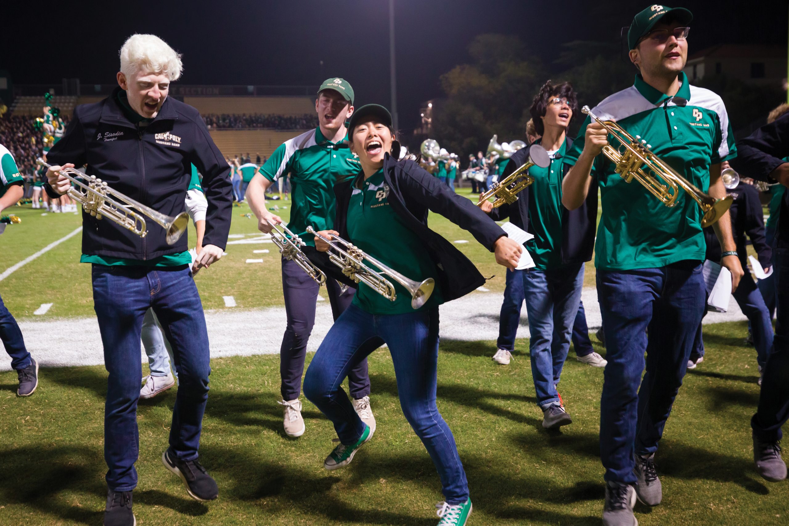 A group of trumpet players in caps and green Mustang Band polos dance wildly on the edge of the football field at Spanos Stadium