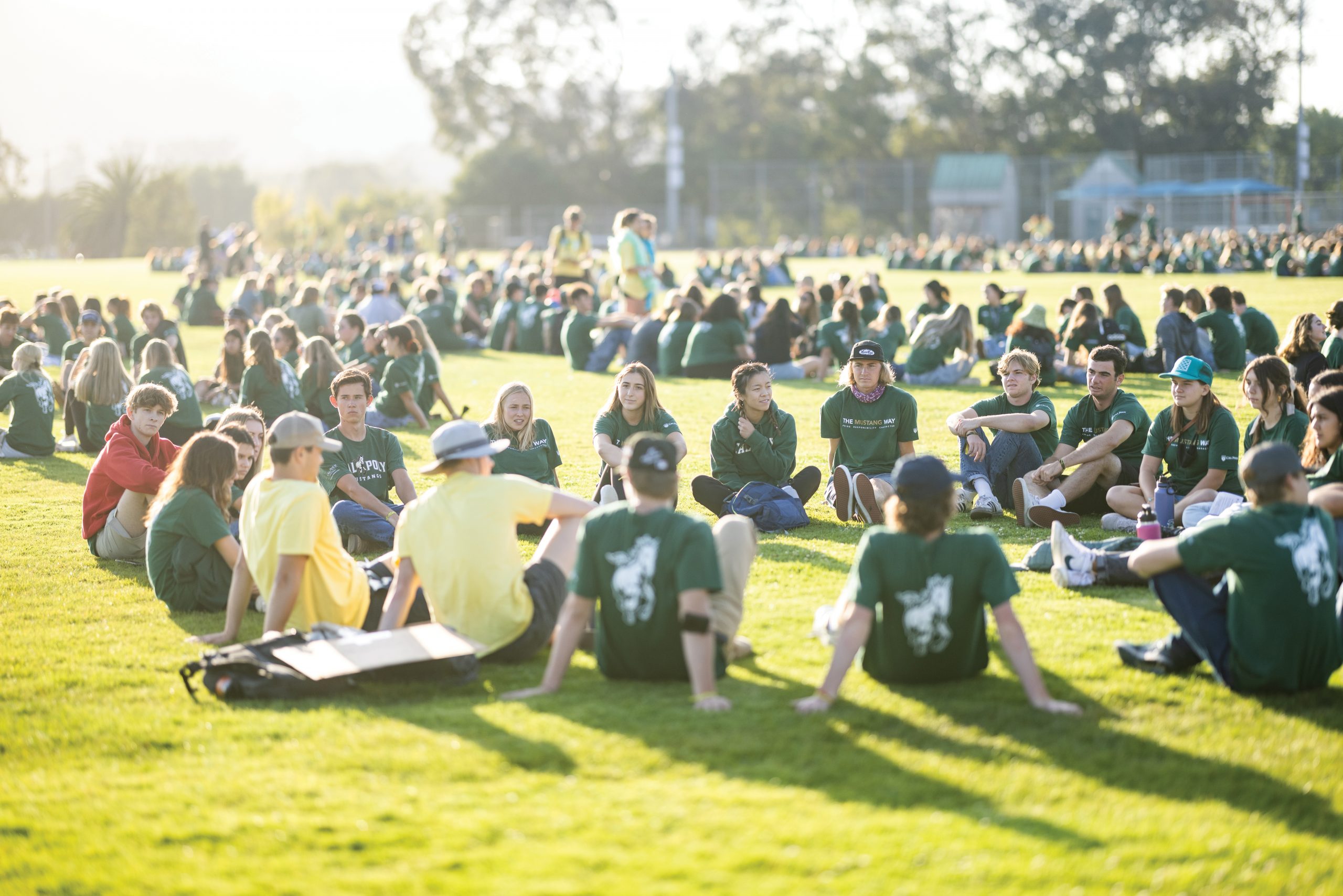 Groups of students in green WOW t-shirts gather in large circles on the grass as the sun sets behind them.