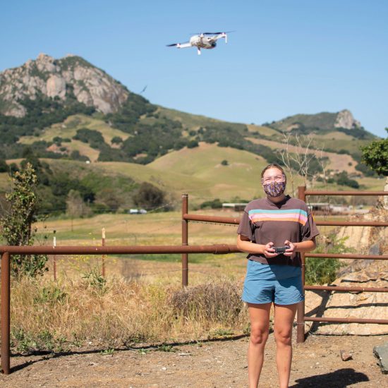 A student controls a drone flying above her head in front of Bishop Peak
