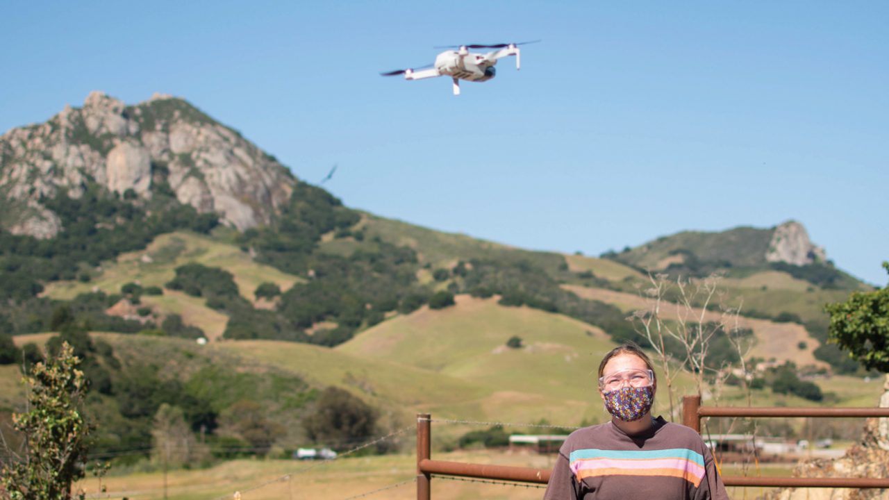 A drone flies above a student standing in front of Bishop Peak