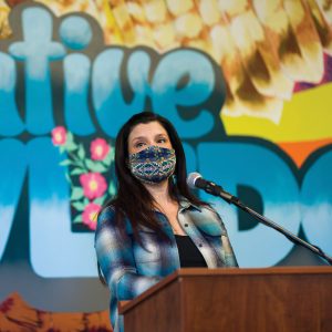 Professor Jenelle Navarro wears a mask while speaking in front of a colorful mural