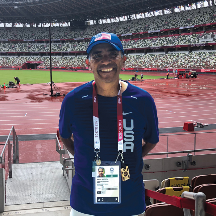 A man wearing Team USA shirt and hat stands in the Tokyo Olympic Track facility wearing a coach's badge