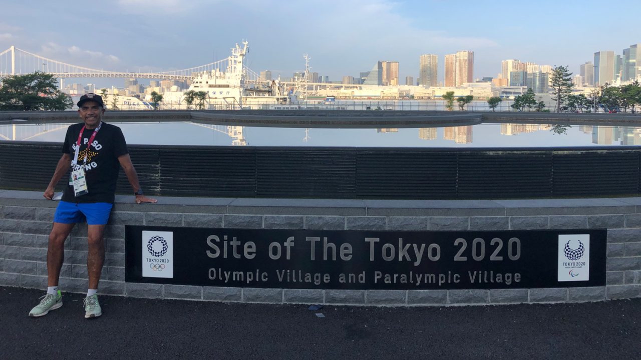 A man stands to the right of a sign in Tokyo saying 'Site of The Tokyo 2020 Olympic Village and Paralympic Village'
