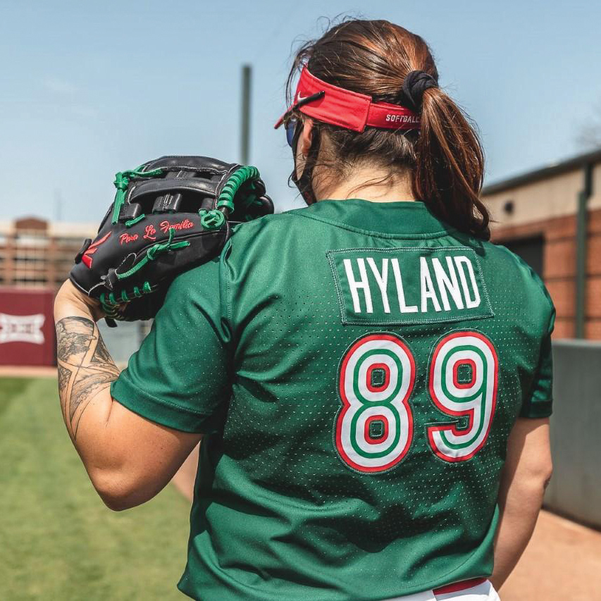 A softball player wearing a mask stands with their back turned, their jersey reading 'Hyland 89'