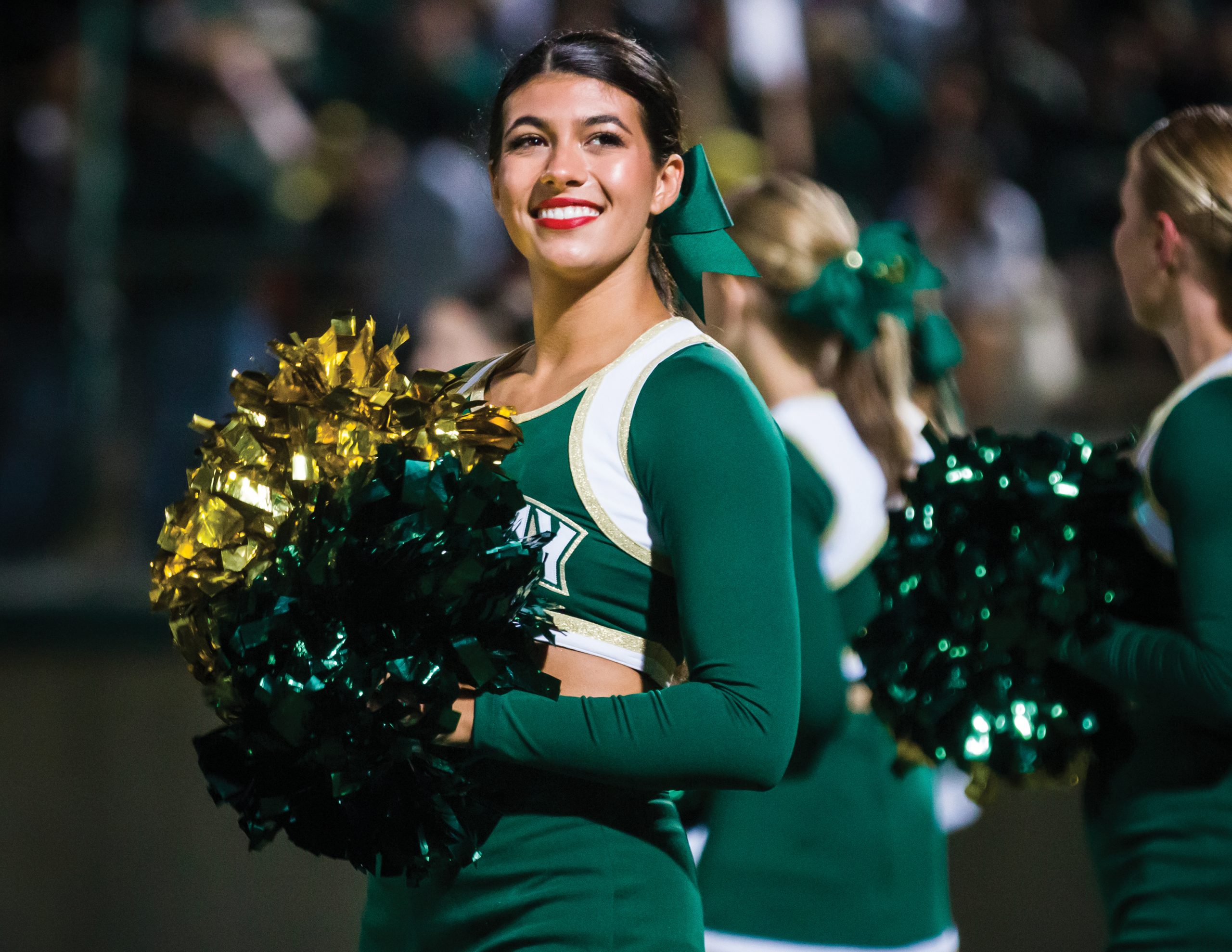 A young woman in a ponytail and green Cal Poly cheerleader uniform holds pom poms and smiles at the crowd in Spanos Statdium