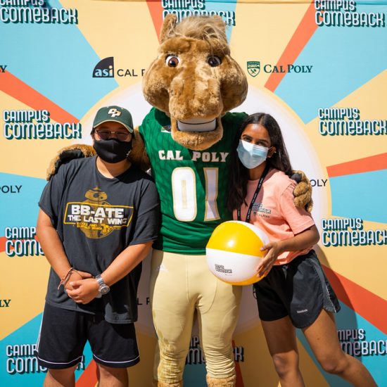 Two people wearing masks hug the Musty the Mustang mascot in front of a colorful backdrop