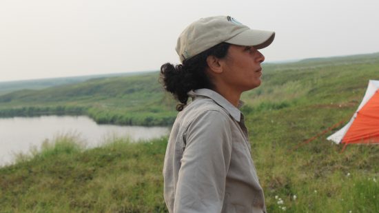 A person wearing a hat and khaki shirt stands in a green field looking to the right