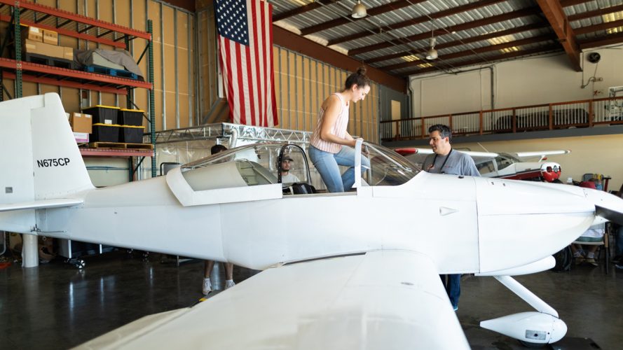 A student climbs into the cockpit of a white airplane in a hangar with an American flag on the wall