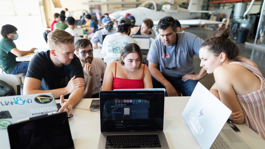 Four students and a professor crowd around a laptop in a hangar with more students and an airplane behind them