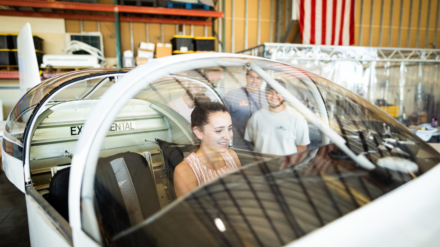 A female student sits in the cockpit of a small, white airplane while a group of students and faculty look on