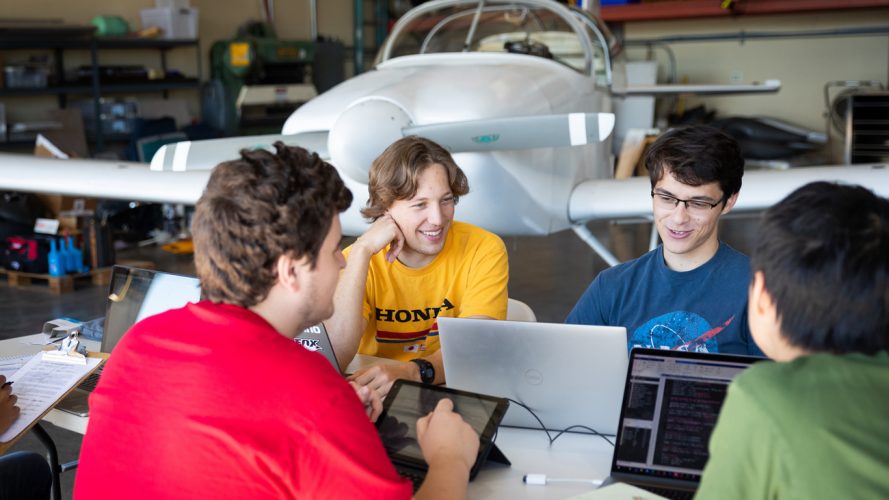 Four students in colorful shirts sit smile while sitting around a laptop in front of a white airplane