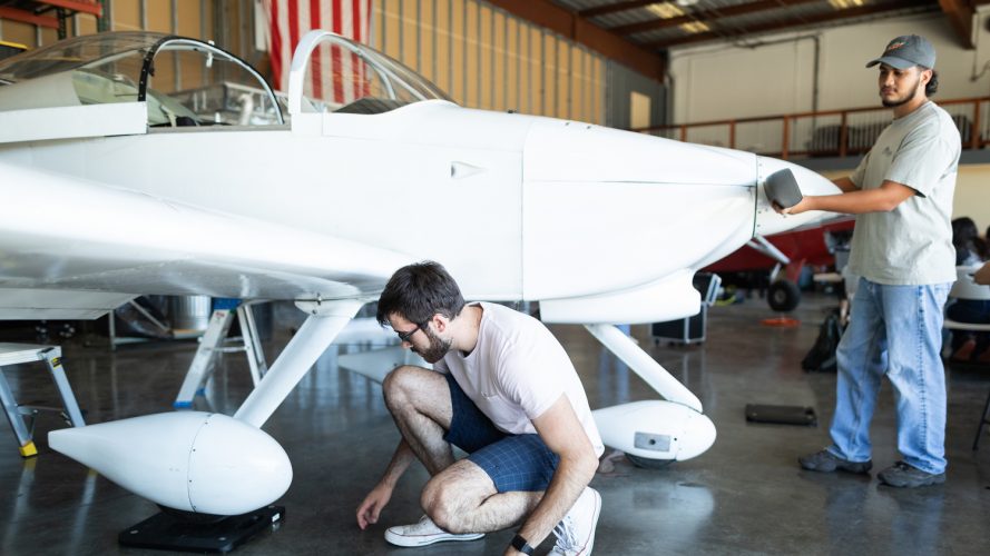 Two students examine a white airplane in a hangar with an American flag in the background