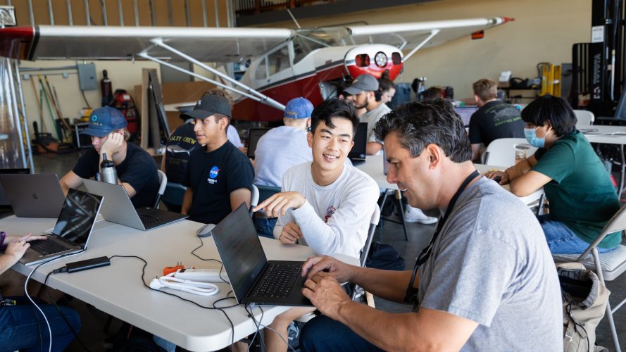 A student and professor look at a laptop in a hangar with a dozen more students and a small airplane