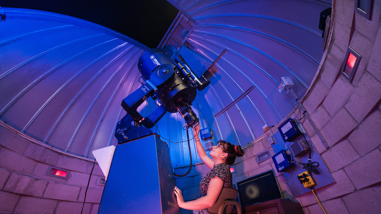 A student smiles as she looks up while handling a large research telescope in an observatory with the roof open to the night sky