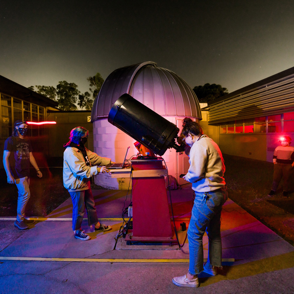Four students stand outside a small cinderblock observatory with one student looking through a telescope