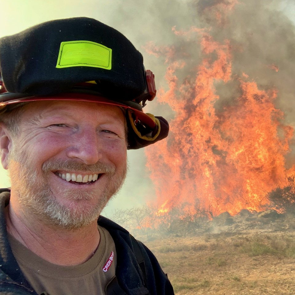 A man in a fire helmet smiles into the camera with flames consuming brush in the bakground